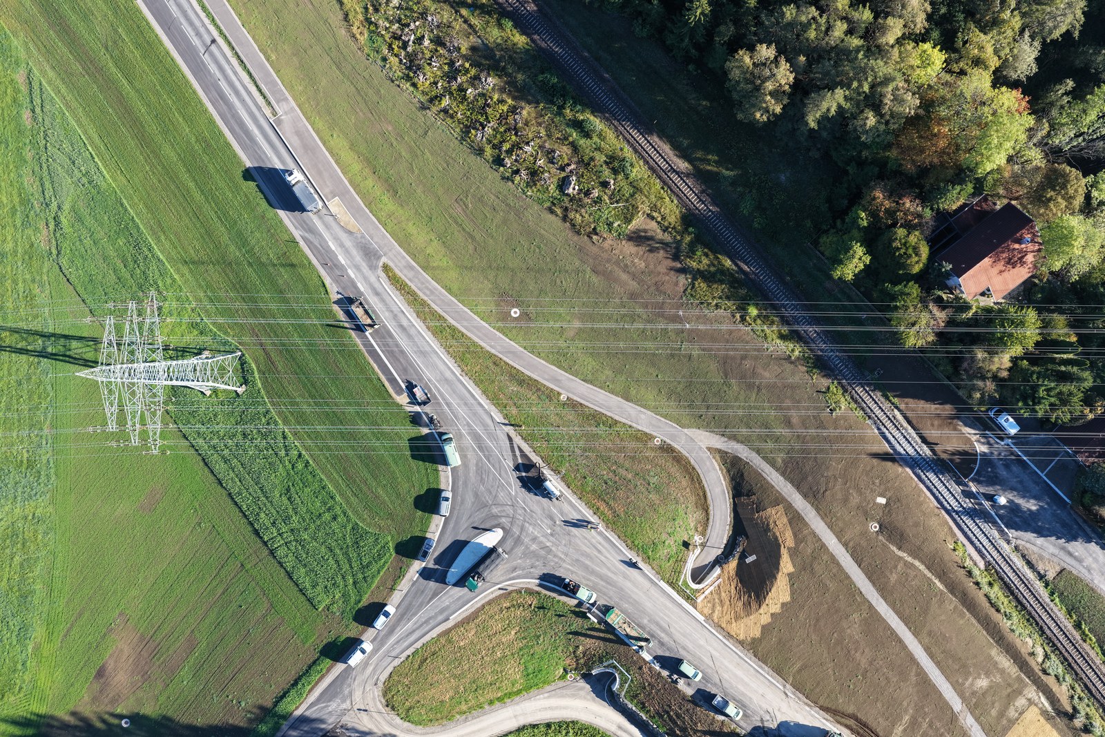 Drohnenaufnahme einer Straße und Bahnstrecke zwischen Feldern und Wald mit einem Strommast.